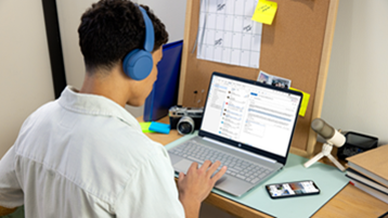 Student wearing blue headphones working on a laptop at a desk with a smartphone, microphone, and camera nearby.